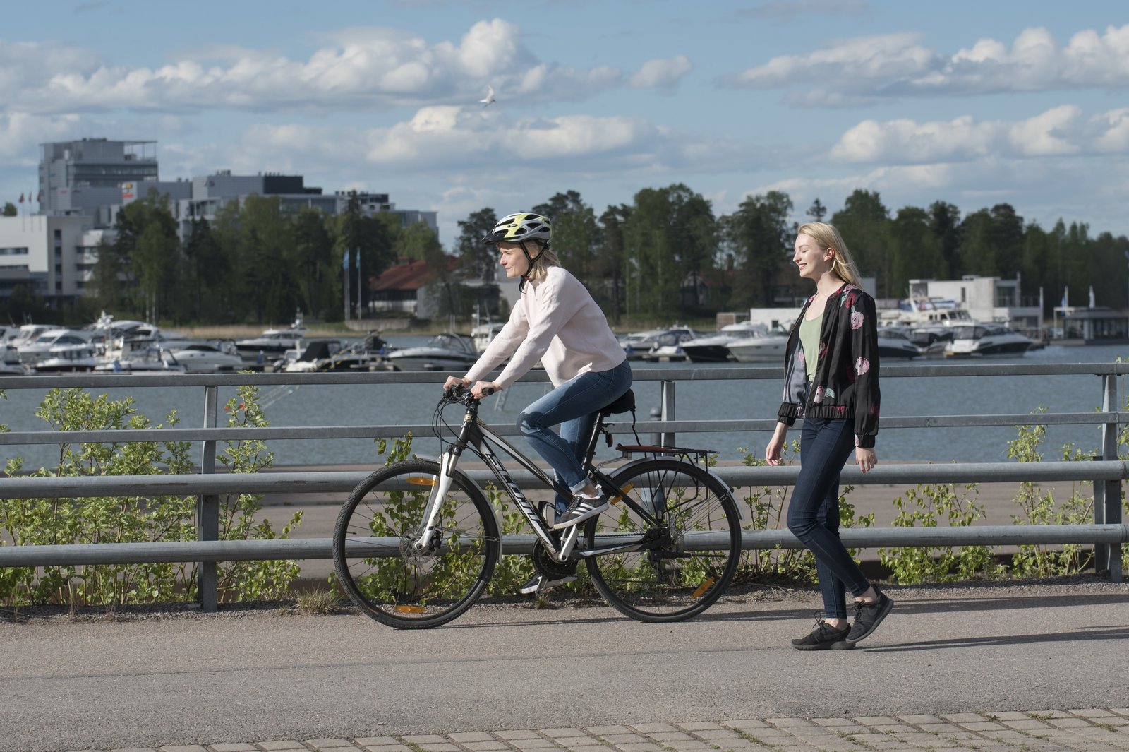 Kaksi nuorta aikuista lenkillä rantaraitilla, toinen pyörällä ja toinen kävellen.