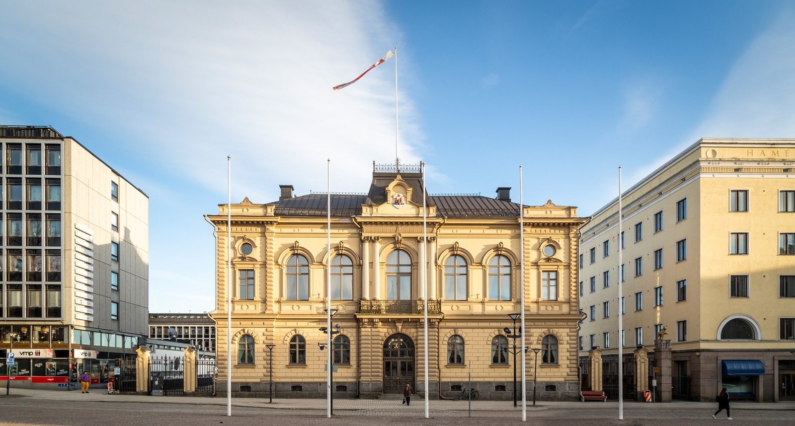 Vaalea rakennus, jonka kummallakin puolella kerrostalot. Taustalla sininen taivas.