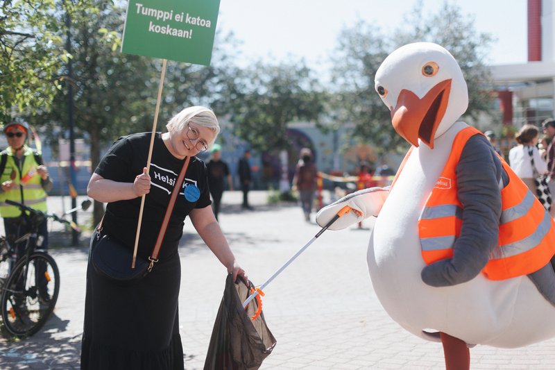 Lokkimaskotti, jolla on päällään heijastinliivi, laittaa roskapihdeillä roskan muovipussiin, jota pitelee vaaleahiuksinen henkilö. Hänen paidassaan lukee Helsinki ja hän pitelee kylttiä, jossa lukee Tumppi ei katoa koskaan!.