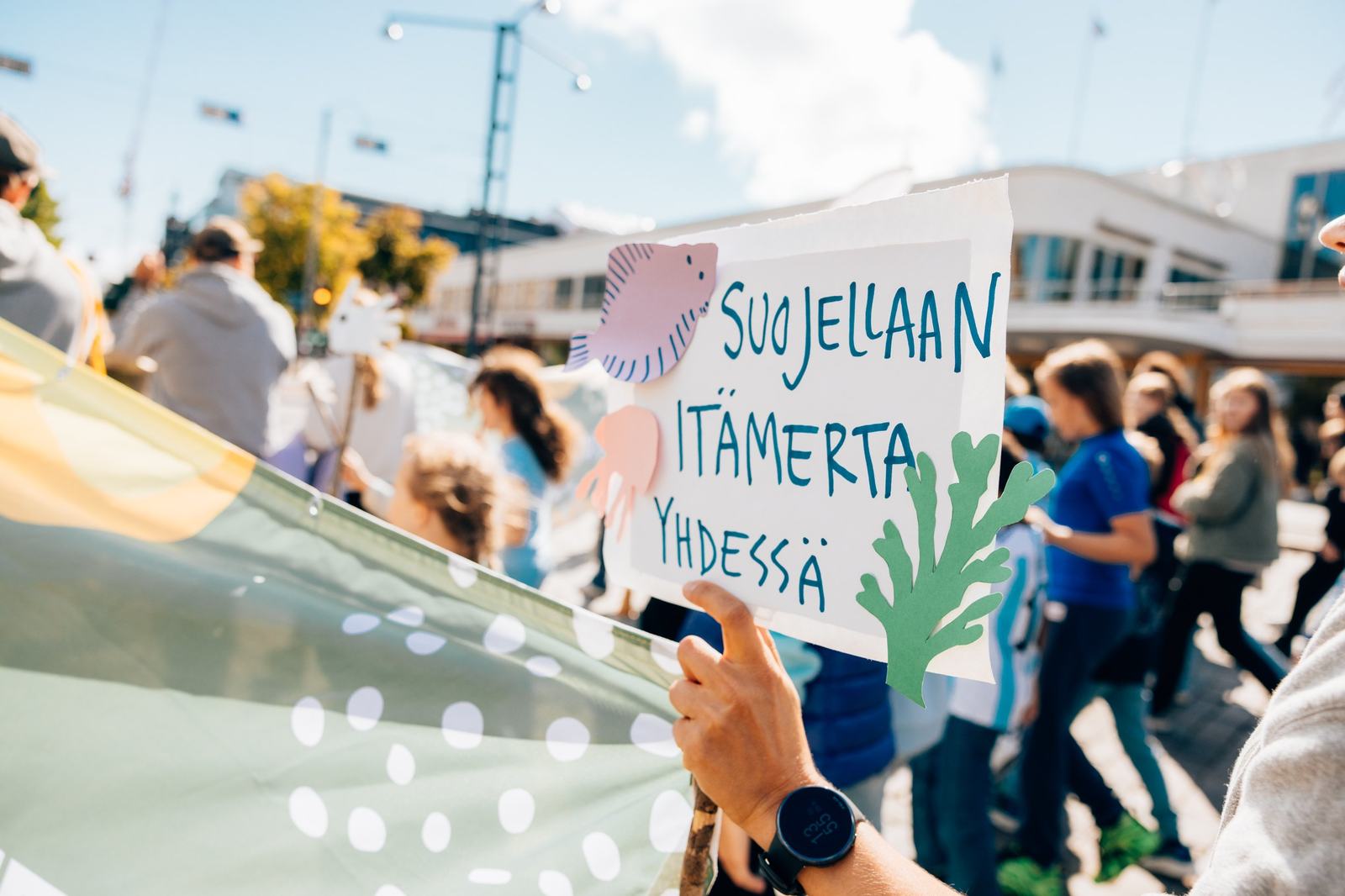 Väkijoukossa ihmishahmoja. Itsetehty kyltti, jossa lukee suojellaan Itämerta yhdessä.